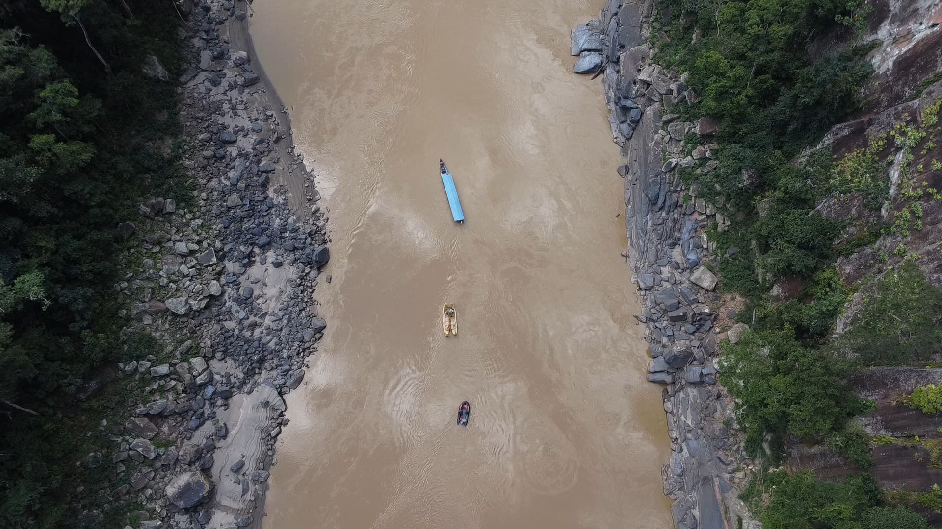 Vue aérienne à la verticale du fleuve brun entre deux rives rocheuses, la balsa suivie de deux embarcations